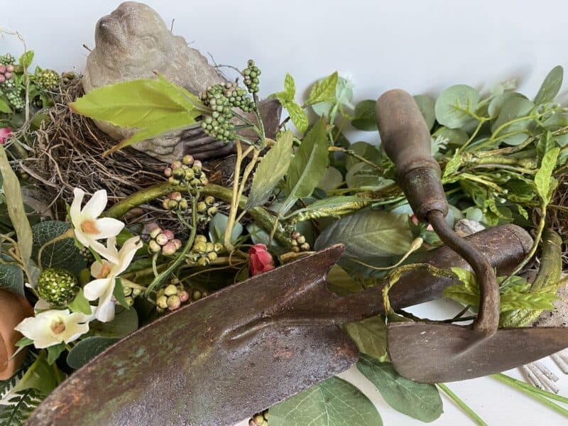 A rustic arrangement featuring a stone bird in a nest surrounded by green leaves, white and pink flowers, and berries. Two vintage garden tools with wooden handles lie in the foreground on the leaves.