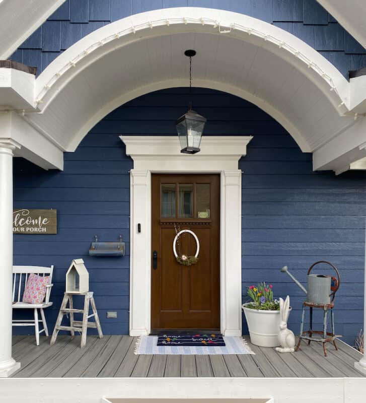 Portion of my front porch which shows the amzing domed roof, brown wood front door with white white trim. There is a pot with spring bulbs on the right was well as a wooden rabbit, a vintage step stool with a vintage water can on the seat. To the left of the door is a portion of a white bench, a small two step ladder that has been white-washed with an IKEA lantern sitting on the top of the step-ladder.