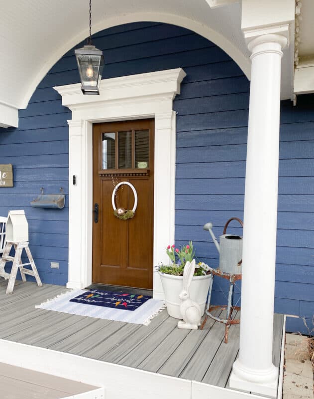 Portion of my front porch which shows the amzing domed roof, brown wood front door with white white trim. There is a pot with spring bulbs on the right was well as a wooden rabbit, a vintage step stool with a vintage water can on the seat. To the left of the door is a portion of a white bench, a small two step ladder that has been white-washed with an IKEA lantern sitting on the top of the step-ladder. Viewed from the side