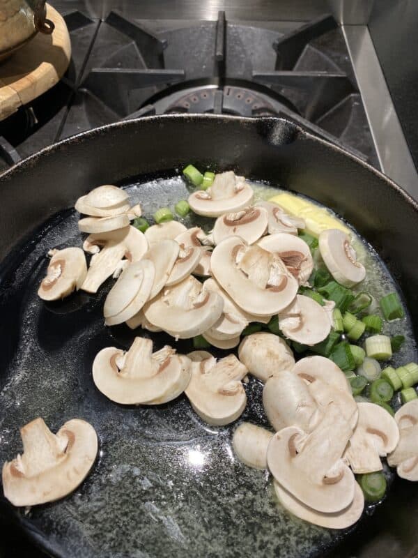 a cast-iron pan on the stove with butter, green onions and mushrooms sautéing