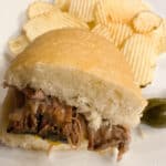 a 1/2 of a beef sandwich with potato chips on a white plate with pickles on the side. This is such an easy slow cooker beef sandwich recipe