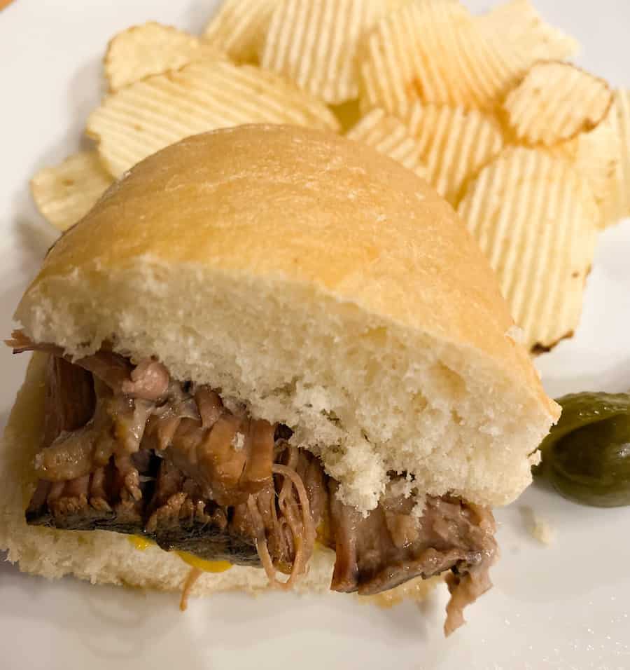 a 1/2 of a beef sandwich with potato chips on a white plate with pickles on the side. This is such an easy slow cooker beef sandwich recipe