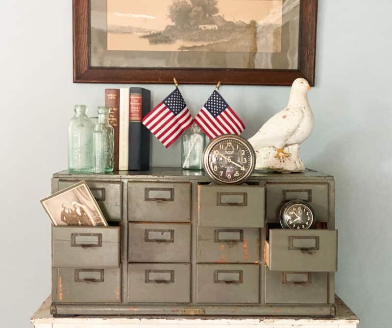 an army green vintage card catalog with some vintage clocks, vintage books, small American flags and a white dove decorating it.