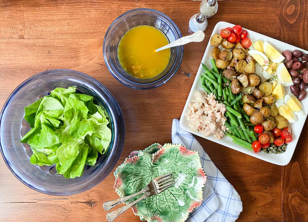 A white platter with the fixings for salad niçoise. A blue vintage bowl filled with bibb lettuce and a smaller blue vintage bowl with the homemade vinaigrette. Two leaf shaped salad plates with two forks.