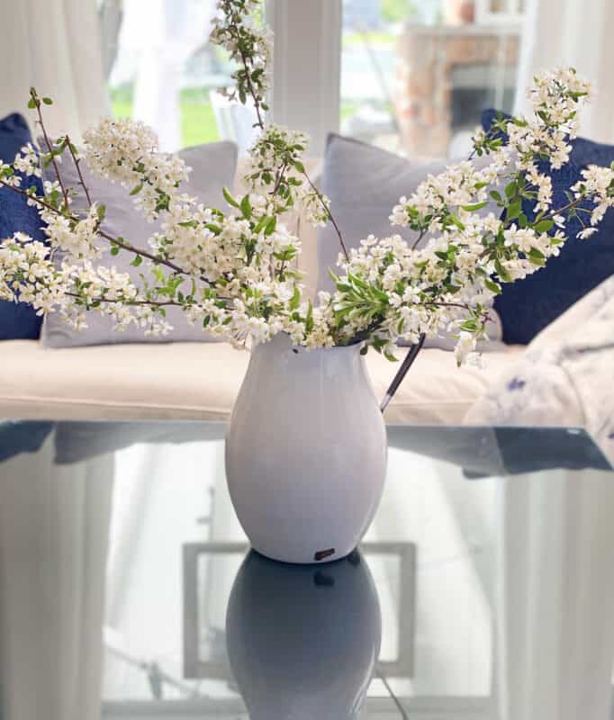 White enamel pitcher with spring branches on a coffee table in front of a white sofa with blue pillows on it. Simple blue and white patriotic decor