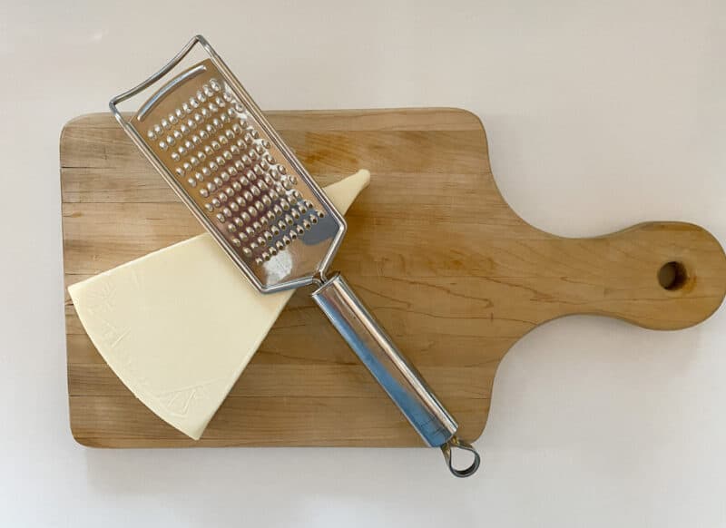 a picture of a cutting board with a block of cheese and a stainless steel grater which is a tool that I use and one of my 10 kitchen shortcuts to make food prep easier