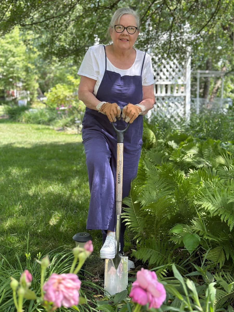 An older woman in a summer capsule wardrobe—white shirt, blue overalls, and gardening gloves—stands on grass, digging with a shovel among lush green plants and pink flowers. Trees and a white trellis frame the vibrant outdoor scene.