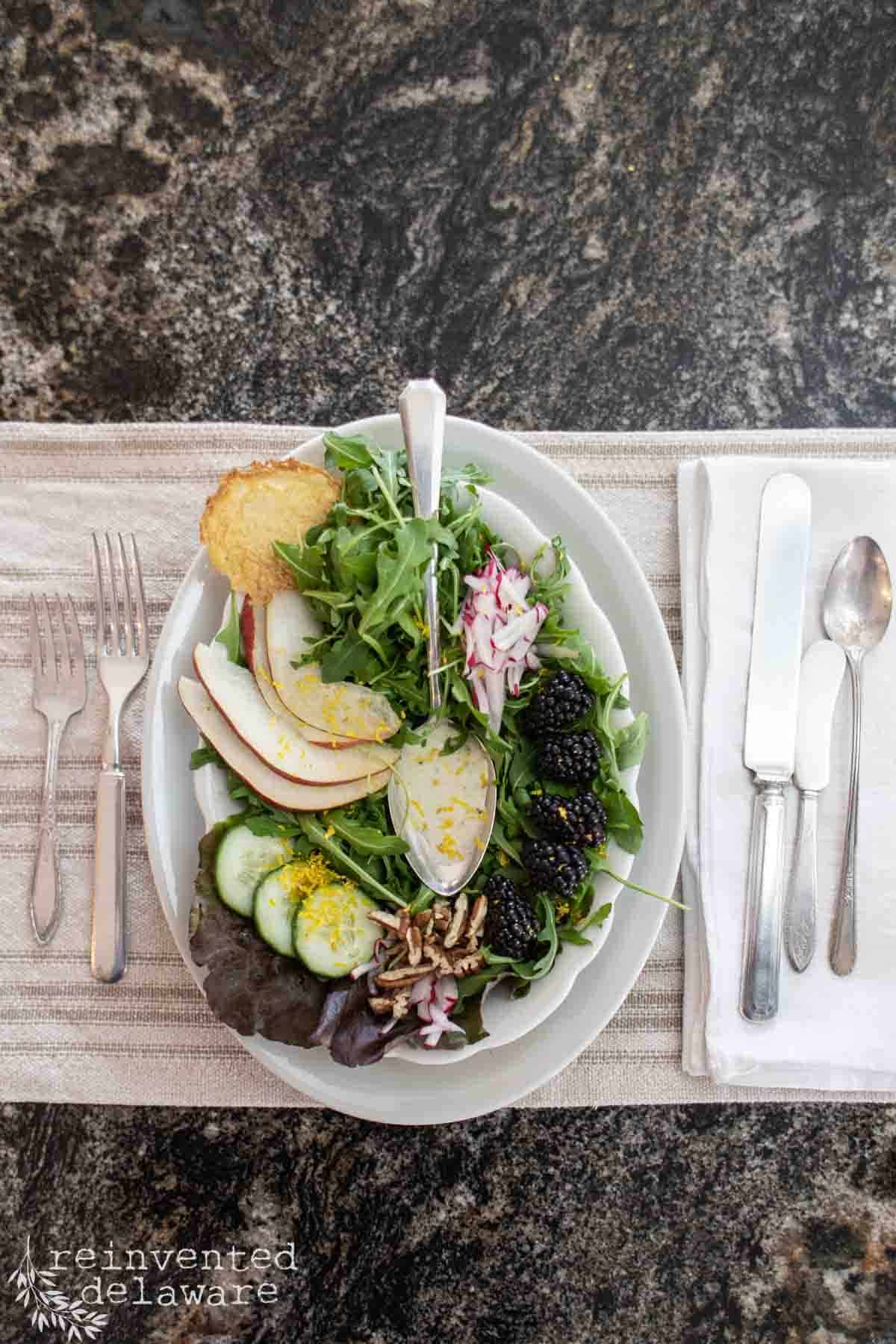 A fresh 4th of July Summer Salad with arugula, sliced pears, cucumber, blackberries, radish, pecans, greens, a cookie, and shredded cheese is served on a white plate with cutlery on a placemat over a dark stone table.
