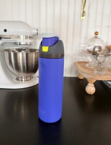 A blue insulated water bottle with a gray and yellow lid sits on a black kitchen counter—primed for kitchen sales—with a stand mixer and glassware in the background.
