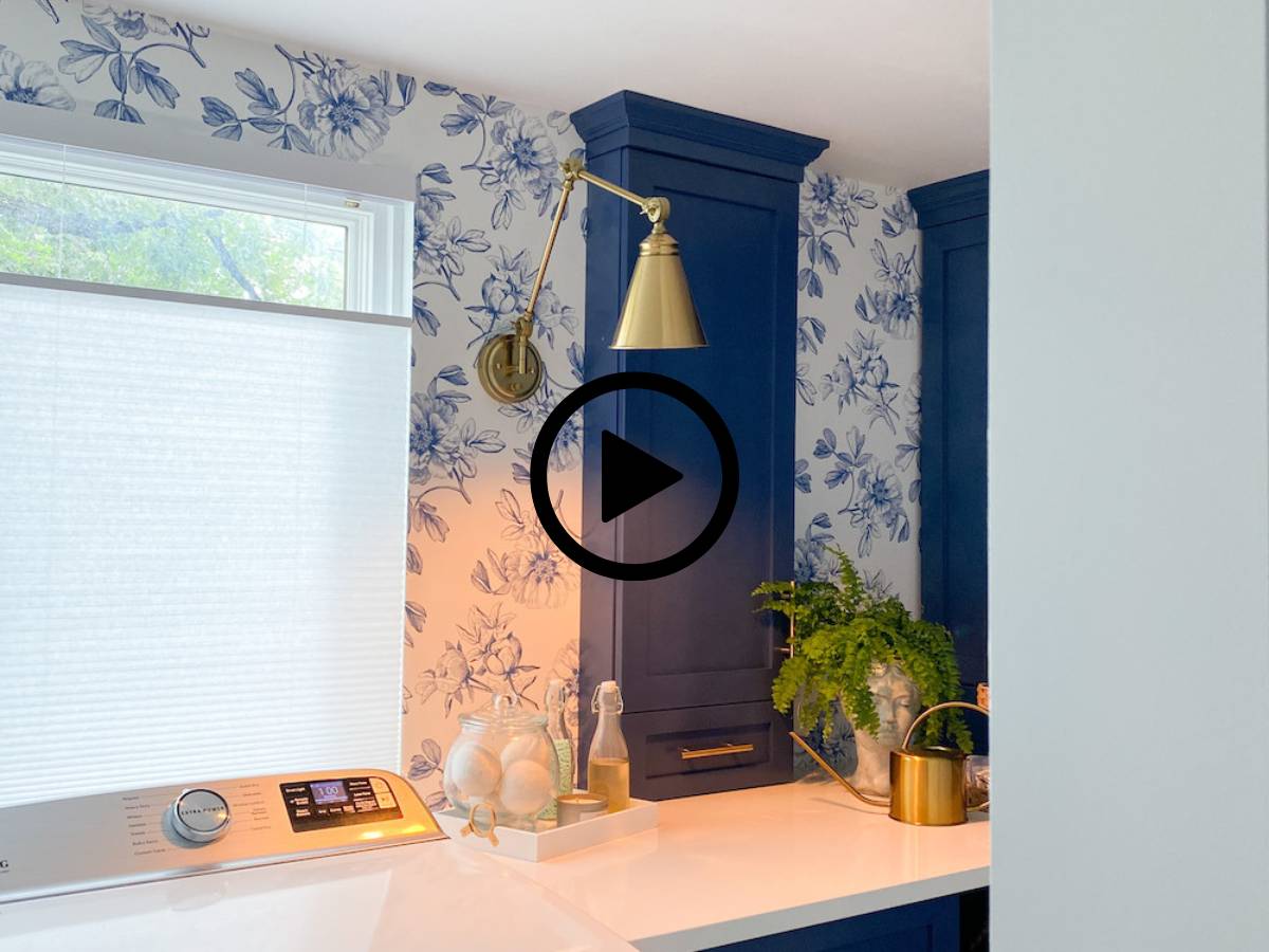 A Small Laundry Room Makeover featuring blue floral wallpaper, blue cabinets, a gold wall sconce, washing machine, white countertop, plant, cleaning supplies, and a gold watering can. A play button icon overlays the center.