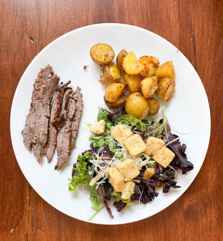 white plate on the table with marinated flank steak, broasted potatoes and a salad
