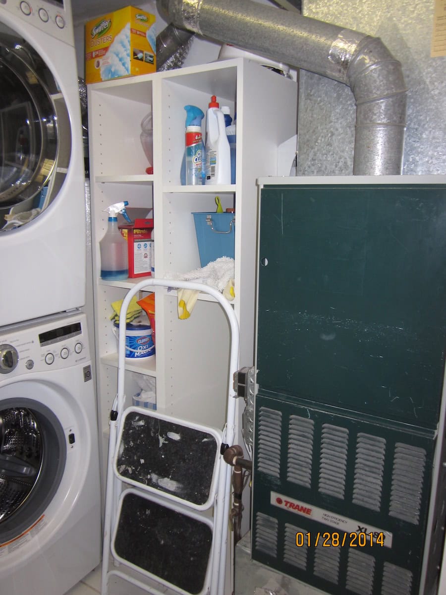 A Small Laundry Room Makeover featuring a stacked washer and dryer, a white shelf with cleaning supplies, a compact folding step ladder, and a green furnace unit with metal ducts.