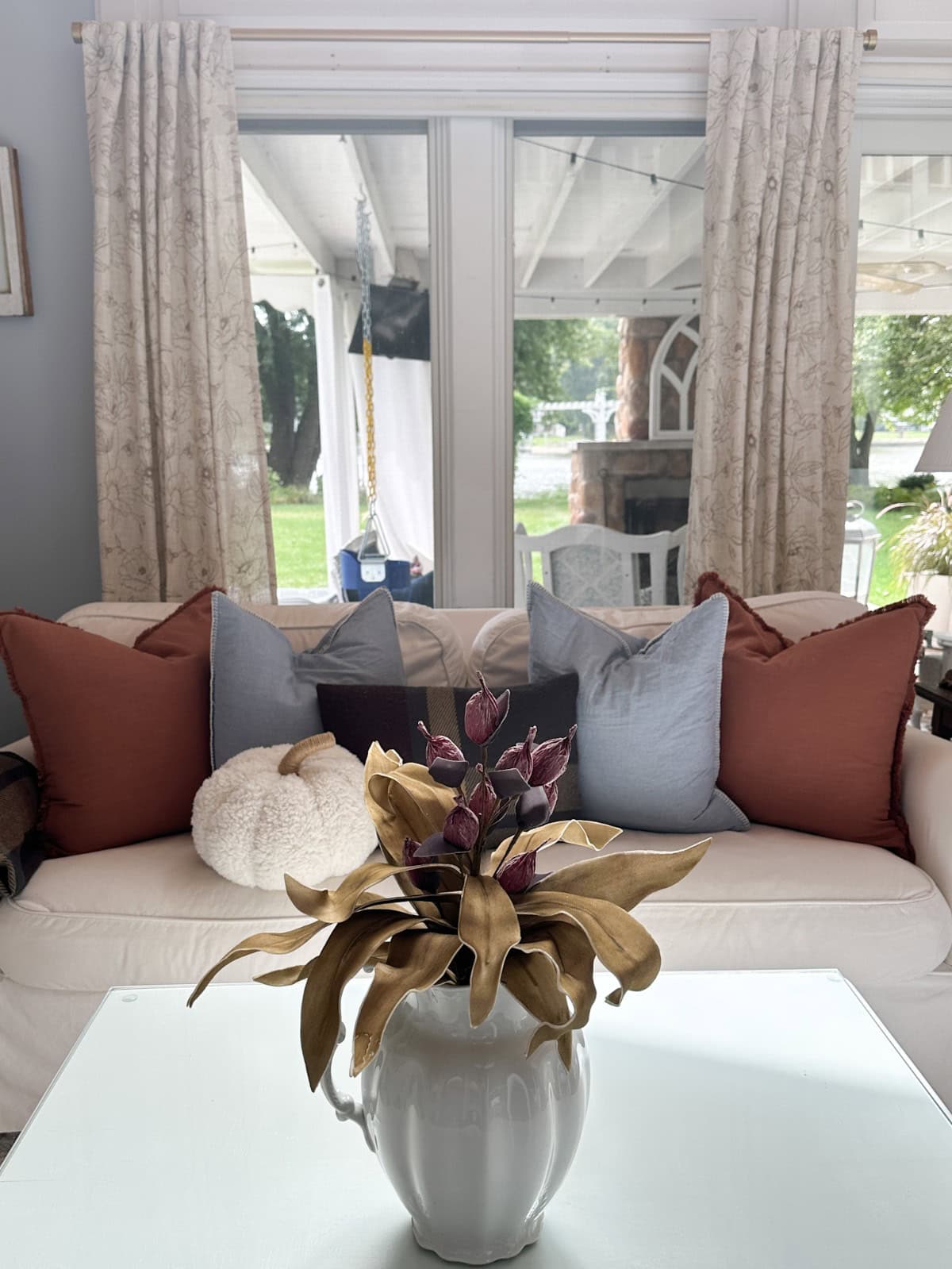 A cozy living room with a white couch adorned with six large pillows in shades of brown, blue, and cream, decorated for the season of autumn. A plush, round cream pillow sits on the left side. In the foreground, a white vase with dried flowers is placed on a glass-top coffee table.