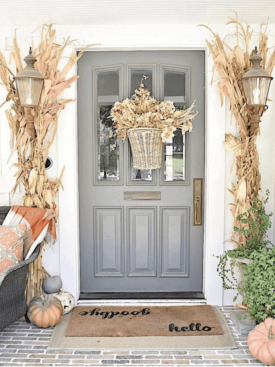 Gray door with Fall Porch Decor: dried flowers, cornstalks, pumpkins, a “hello” mat, and a potted plant nearby.
