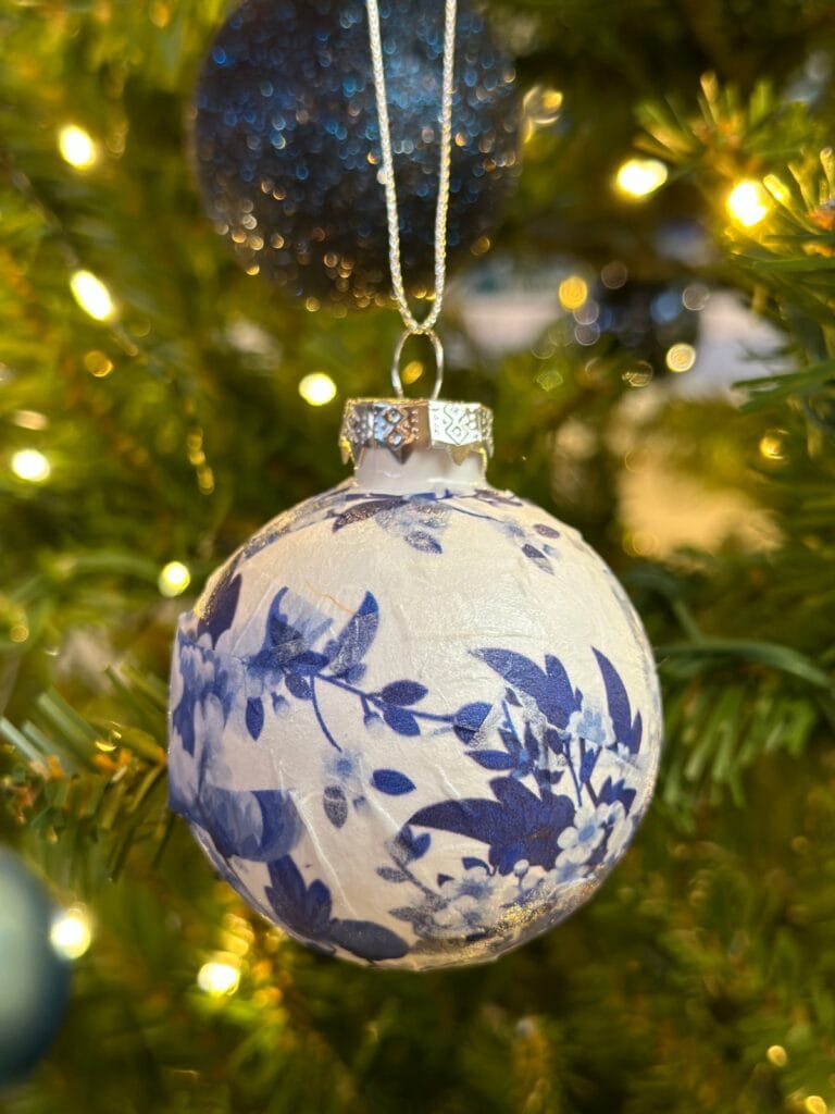 A close-up of a Christmas ornament with blue floral patterns hanging on a lit French Country Christmas Tree, with blurred lights and branches in the background.