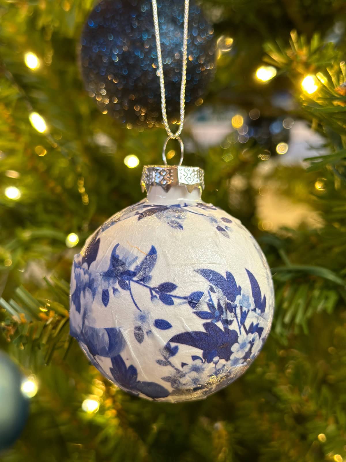 A close-up of a Christmas ornament with blue floral patterns hanging on a lit French Country Christmas Tree, with blurred lights and branches in the background.