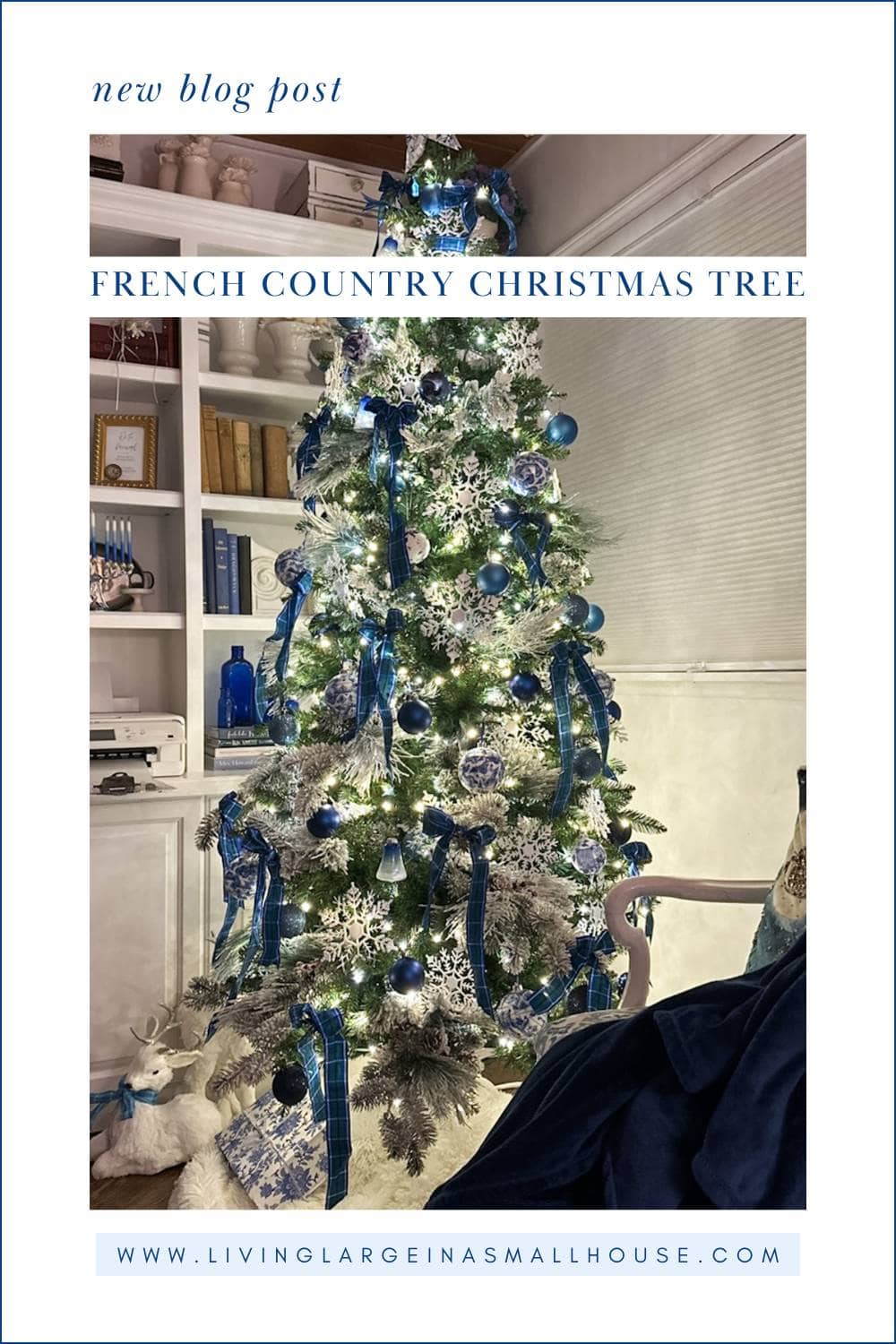 A French Country Christmas Tree adorned with blue ribbons, ornaments, and white snowflakes stands by shelves of books and blue vases. A plush white reindeer sits at the base, inspiring a blog post on elegant French country Christmas decor.