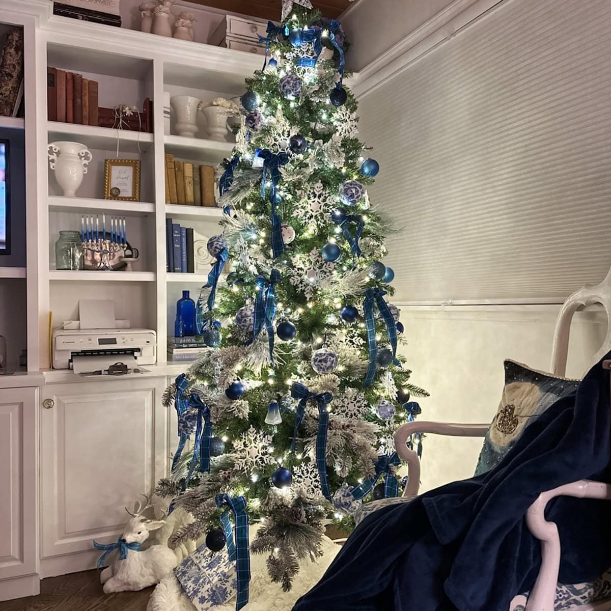A French Country Christmas Tree with blue and white ornaments, ribbons, and lights stands in a cozy living room with bookshelves and holiday decor, featuring a fluffy white tree skirt and a small reindeer figure underneath.