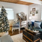 Cozy living room with a French Country Christmas Tree, vintage furniture, a “BELIEVE” pillow on a white chair, an old trunk as a coffee table with mini Christmas trees, and charming holiday decor throughout.