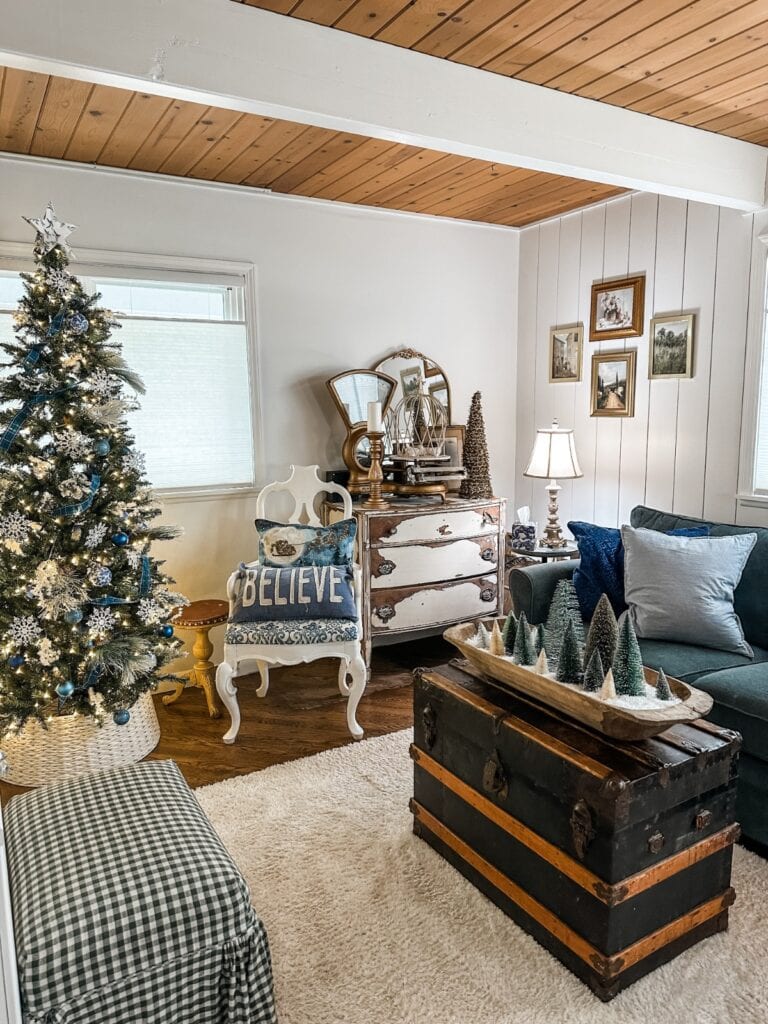 Cozy living room with a French Country Christmas Tree, vintage furniture, a “BELIEVE” pillow on a white chair, an old trunk as a coffee table with mini Christmas trees, and charming holiday decor throughout.
