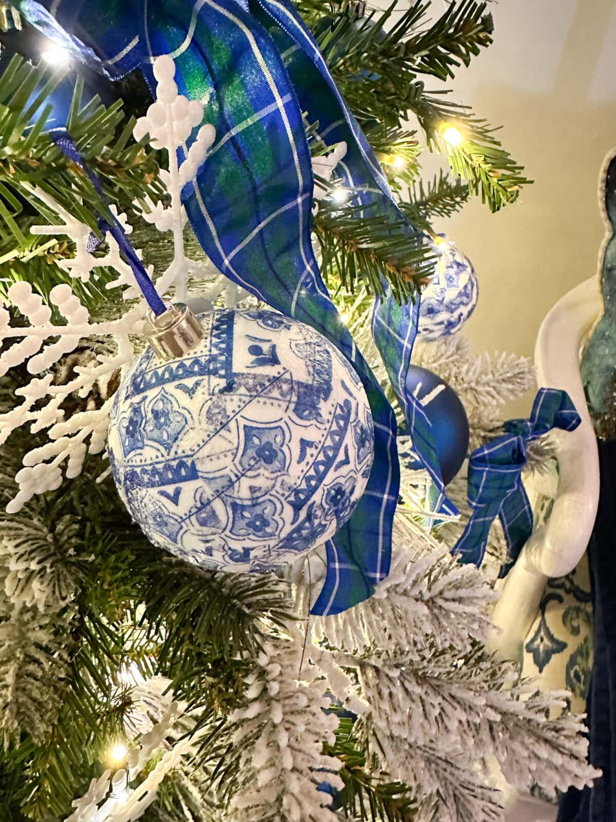A close-up of a French Country Christmas Tree decorated with a blue and white patterned ornament, a blue plaid ribbon, white faux branches, and warm white string lights.