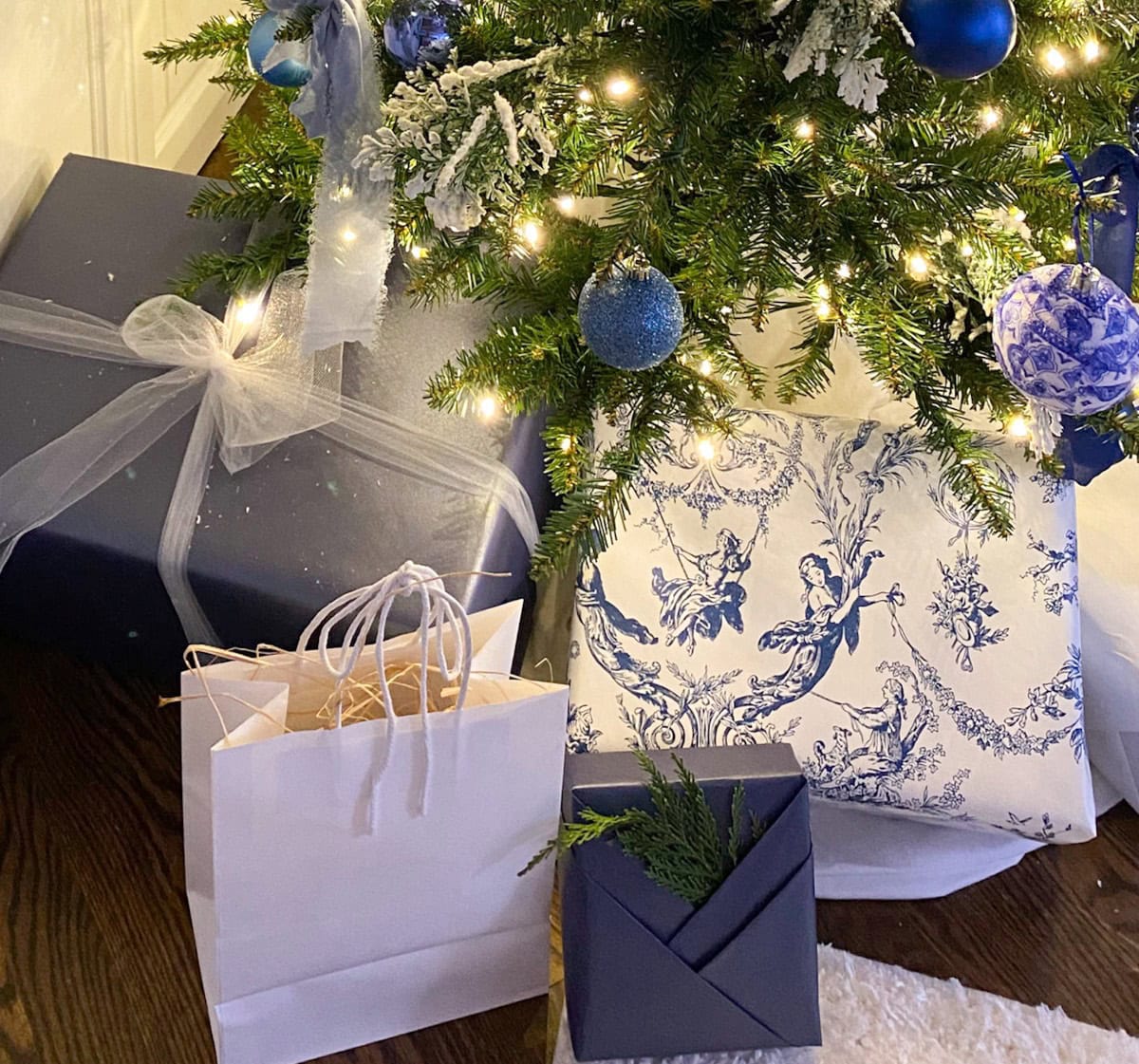 Wrapped gifts in blue, white, and patterned paper sit beneath a French Country Christmas Tree adorned with blue and white ornaments and glowing lights. One white gift bag rests on the wooden floor.