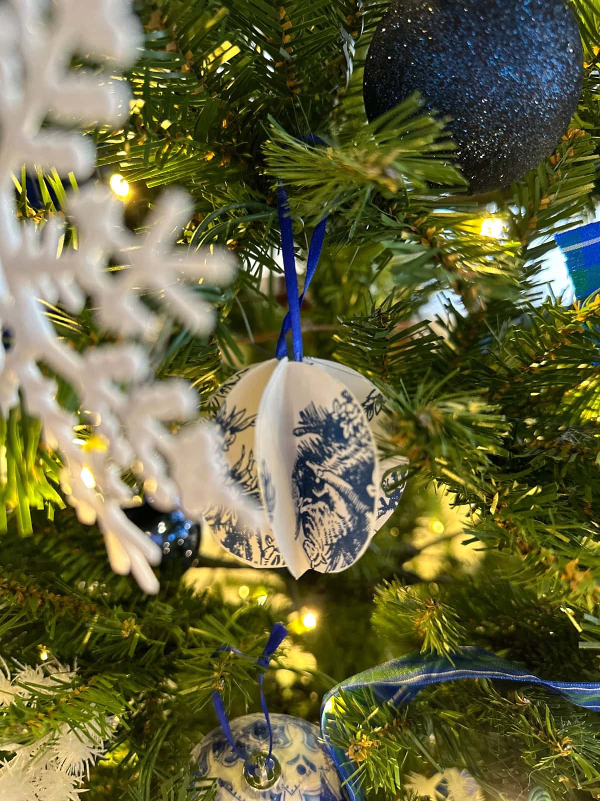A close-up of a French Country Christmas Tree decorated with a blue and white paper ornament, a white snowflake ornament, blue ribbon, and a glittery blue ball ornament, all surrounded by green pine needles and glowing lights.