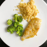 A white plate with a serving of cooked broccoli, brown rice, and a piece of seasoned easy skillet lemon chicken breast on a black surface.