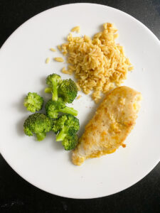 A white plate with a serving of cooked broccoli, brown rice, and a piece of seasoned easy skillet lemon chicken breast on a black surface.