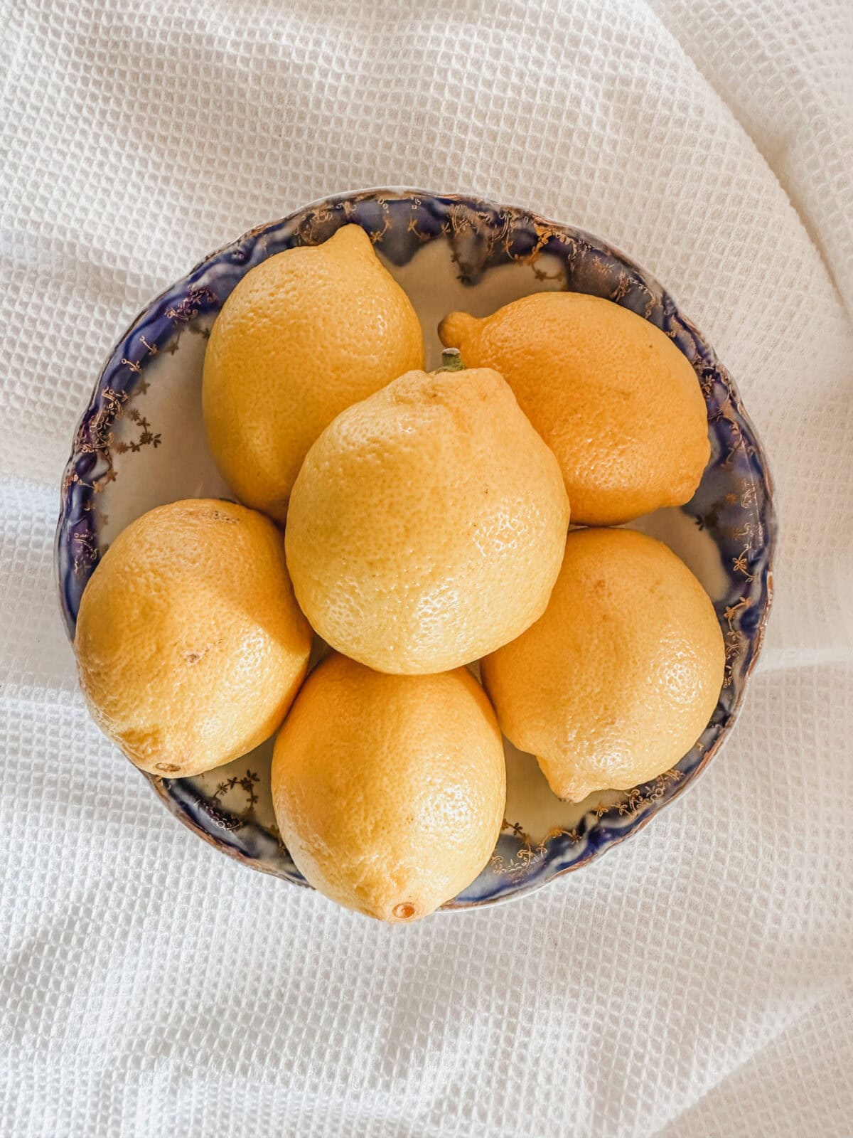 A ceramic bowl with a blue rim holds six yellow lemons, perfect for making Lemon Chicken, placed on a white textured fabric background.