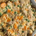 A close-up of a casserole dish filled with a creamy mixture of chicken, peas, carrots, and stuffing—a perfect answer to "What can you make with leftovers from Thanksgiving?" The filling features chunky vegetables and meat in a thick, yellow sauce.