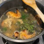 A pot of homemade chicken stock simmering on a stove, made from roasted chicken pieces—perfect for using leftovers from Thanksgiving—with fresh herbs and a wooden spatula resting on the side as steam rises from the bubbling broth.
