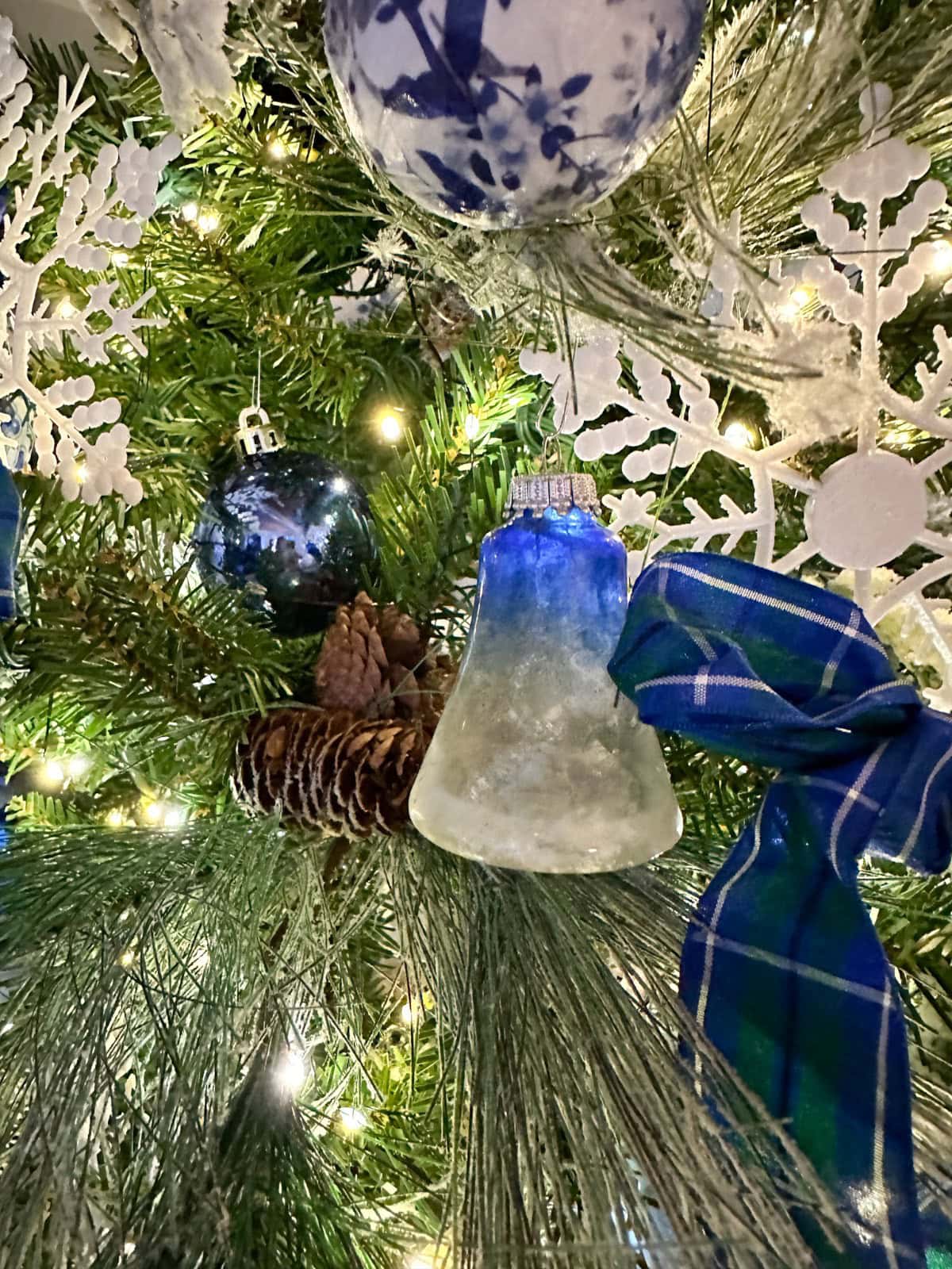 A close-up of a French Country Christmas Tree decorated with a blue-and-white bell ornament, blue ornaments, white snowflake decorations, pinecones, a blue plaid ribbon, and twinkling lights.