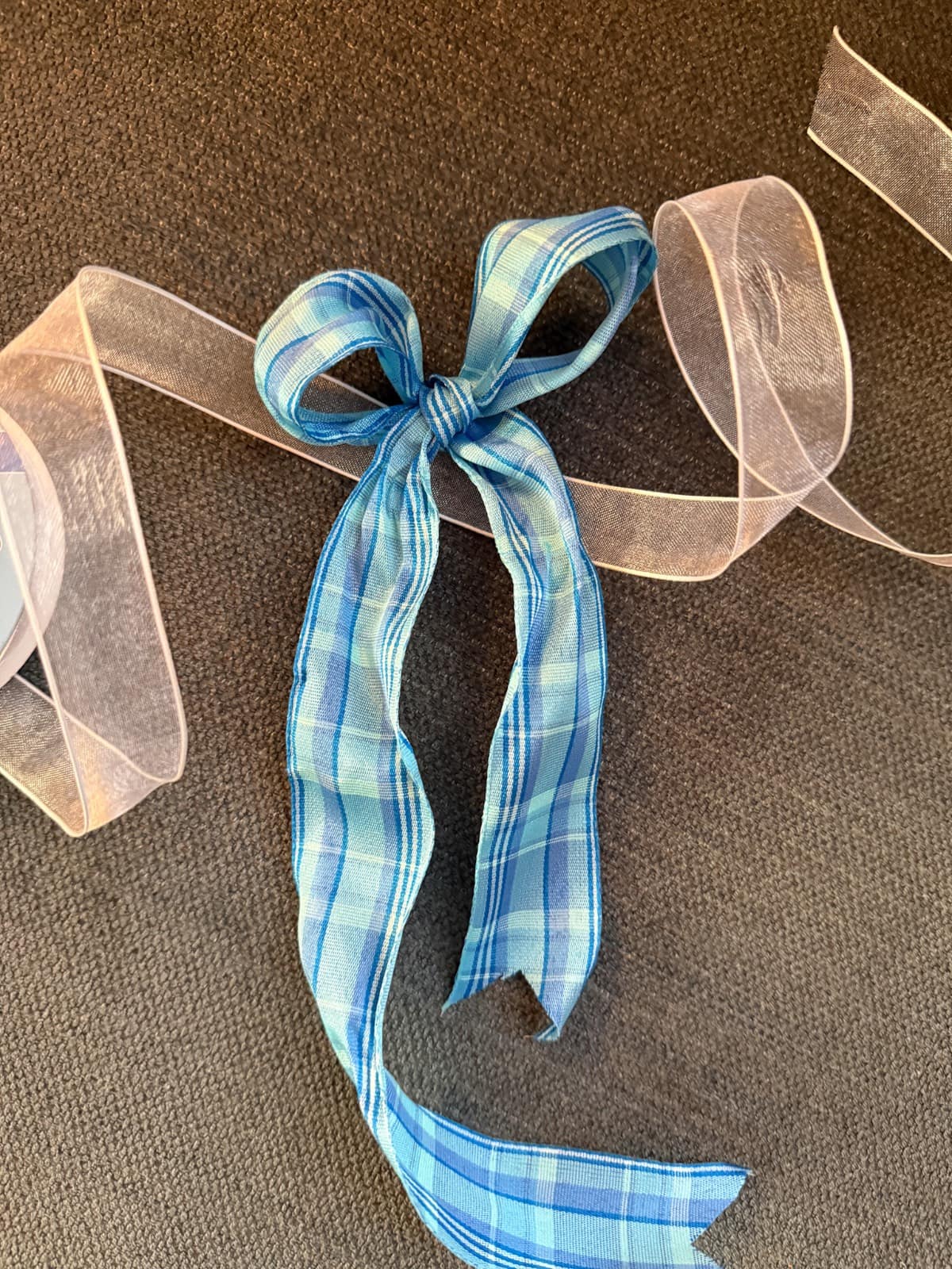 A blue plaid ribbon is tied in a bow on a dark textured surface, with a sheer pink ribbon curling around it—perfect for adding French Country Christmas Tree charm to your holiday décor.