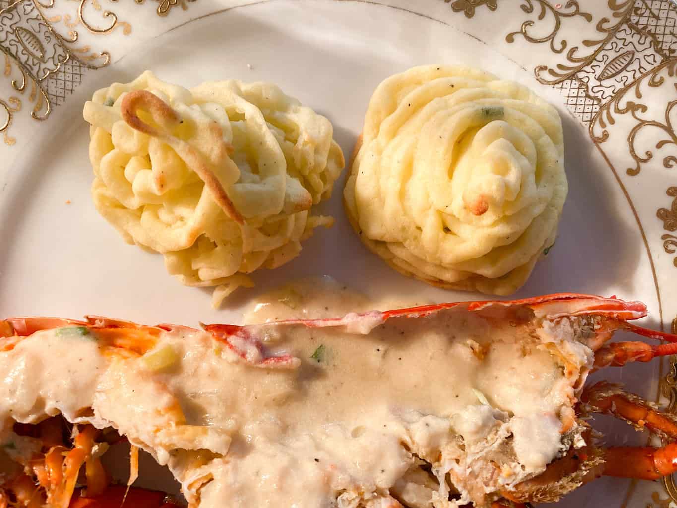 A plate with two swirled servings of mashed potatoes and half a lobster topped with creamy white sauce, reminiscent of dining on the Orient Express, is served on a decorative white and gold-patterned plate.
