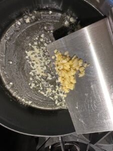 Chopped garlic being added to a pan with sautéing onions in melted butter or oil using a metal bench scraper, forming the aromatic base for Potato Broccoli and Leek Soup over a stovetop.
