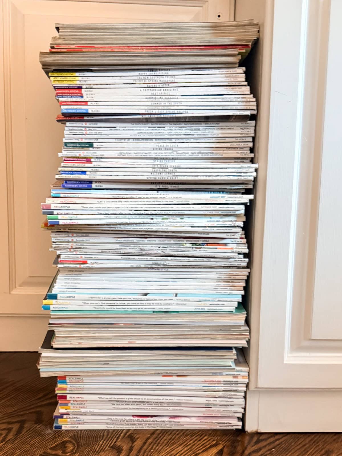 A tall stack of assorted magazines, perfect for organizing my paperwork, sits neatly on the wooden floor by a white cabinet.