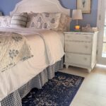 A cozy bedroom to refresh your home for spring, featuring a white bed with floral bedding, a blue gingham bed skirt, and a patterned rug. A white nightstand holds a lamp, candle, books, and framed art on the light blue wall.