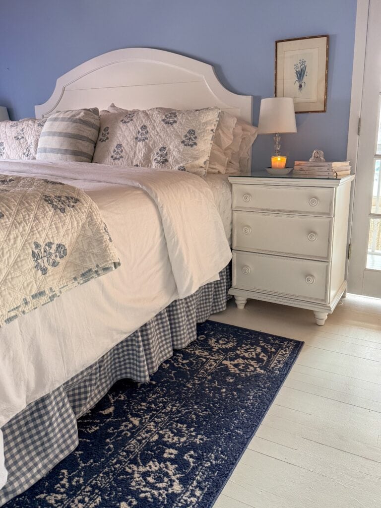 A cozy bedroom to refresh your home for spring, featuring a white bed with floral bedding, a blue gingham bed skirt, and a patterned rug. A white nightstand holds a lamp, candle, books, and framed art on the light blue wall.
