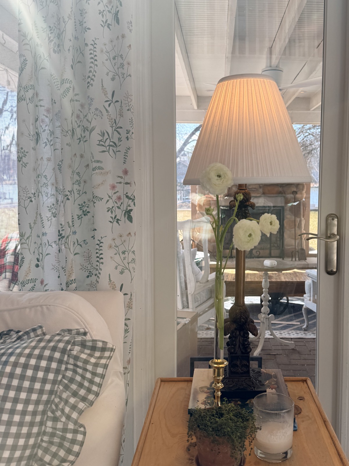 Refresh your home for spring with a cozy indoor scene: a gingham pillow and floral curtain beside a wooden side table holding a lamp, potted plant, candle, and glass of milk. A patio with furniture is visible outside through the glass door.