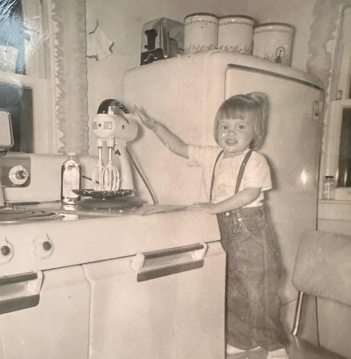 A young child smiling and standing on a chair in a vintage kitchen, reaching toward a stand mixer beside a stove and old-fashioned refrigerator. This black-and-white photo captures joyful recollections of simpler times.