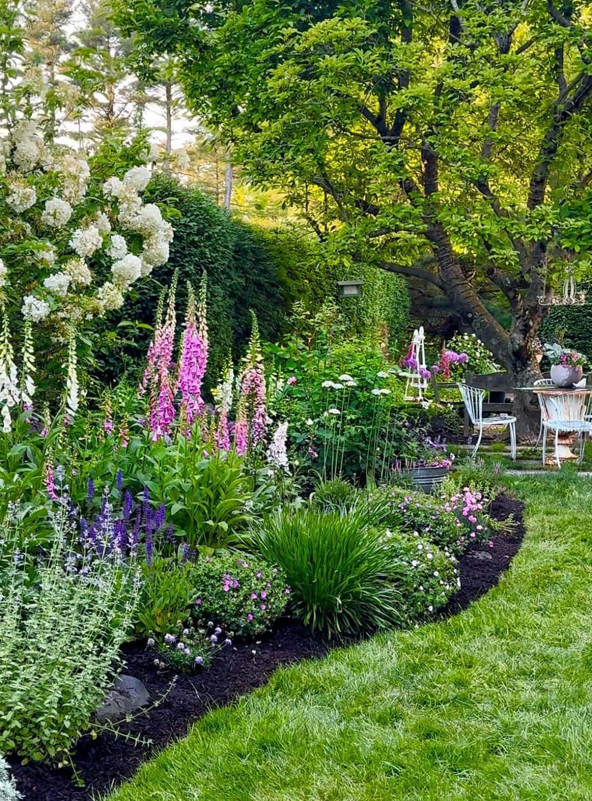 A lush farmhouse cottage garden with blooming flowers in shades of pink, white, and purple runs alongside a green lawn. Outdoor seating beneath large leafy trees in the background creates a peaceful, inviting atmosphere.