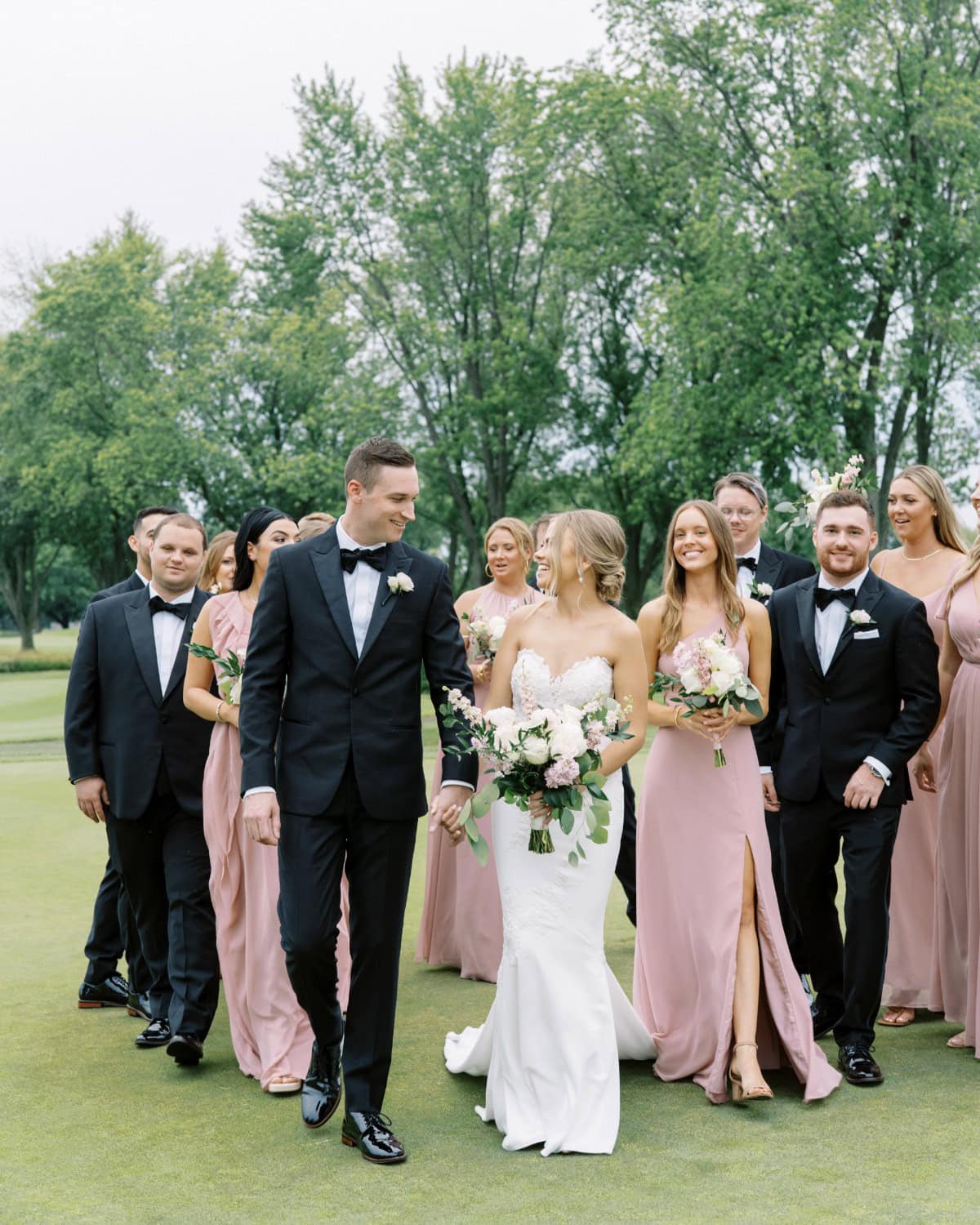 A bride and groom walk hand-in-hand outdoors at their intimate vintage backyard wedding, smiling and surrounded by cheerful bridesmaids in mauve dresses and groomsmen in black tuxedos. Lush green trees and grass complete the charming scene.