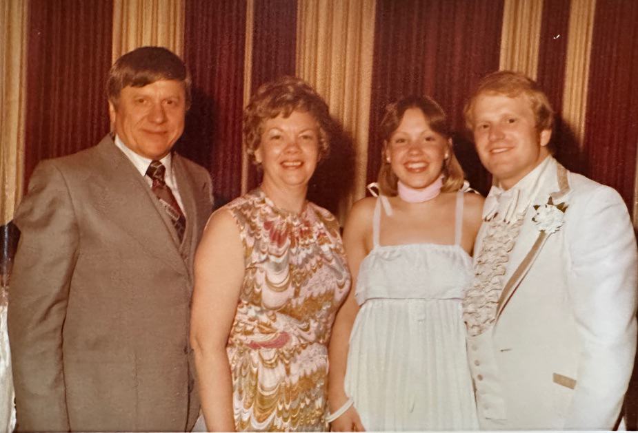 Four people pose together in formal 1970s attire, a scene full of recollection. Two men wear suits—one with a tie, the other with a boutonnière—while two women in patterned and white dresses smile before striped curtains.