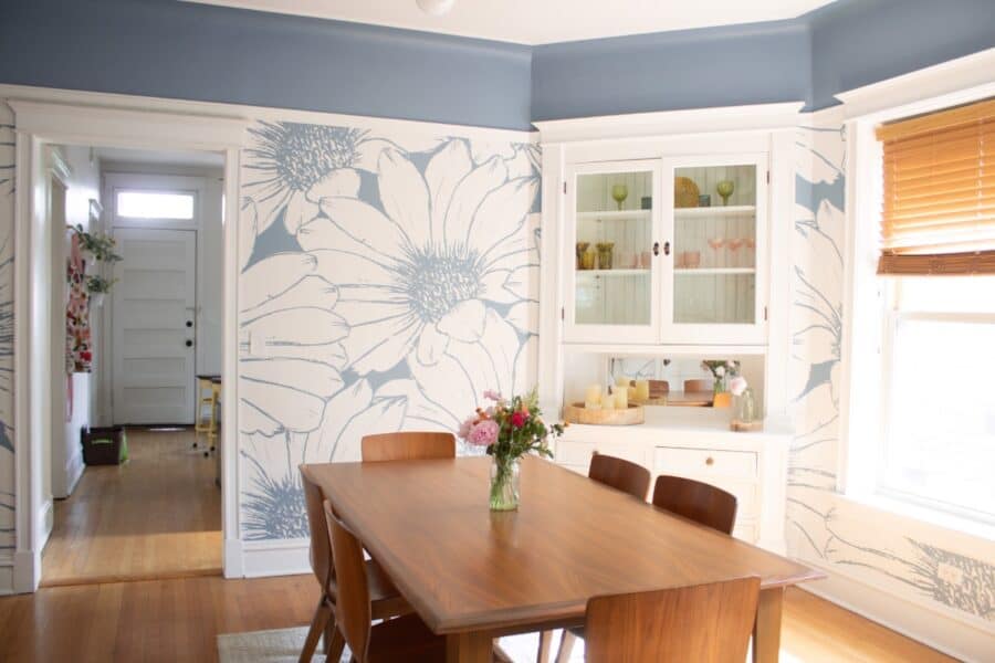 A bright dining room with a wooden table and six chairs, blue and white peel and stick wallpaper, a china cabinet, and a window with wooden blinds letting in natural light. A vase with flowers sits on the table.