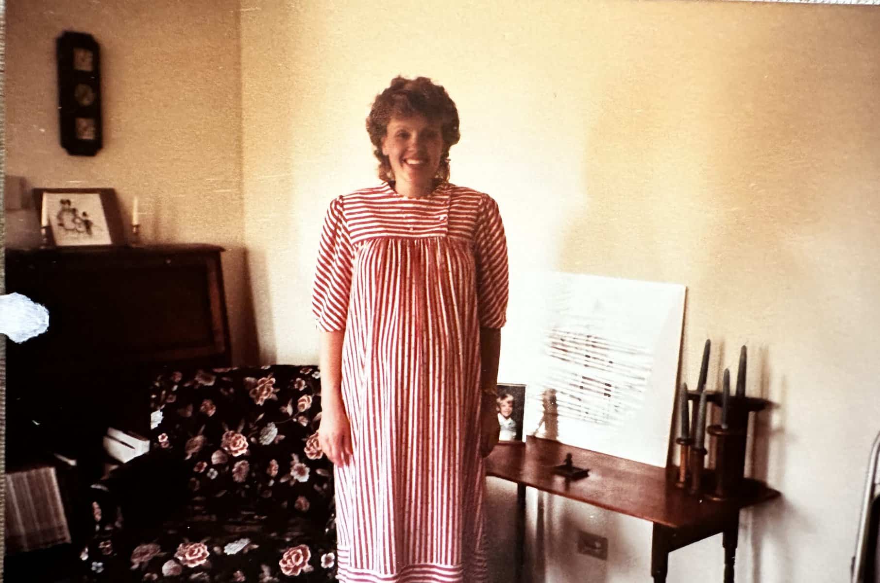 A woman in a red and white striped dress stands smiling in a living room filled with recollection—a floral armchair, small table with candles, a picture frame, and artwork rest against the cream-colored wall.