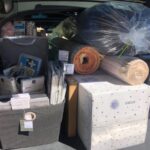 The car trunk is expertly packed with a rolled carpet, black trash bag, storage bins, cardboard boxes, and household supplies—everything from your college dorm checklist. The space is neatly organized, maximizing every inch of the trunk area.