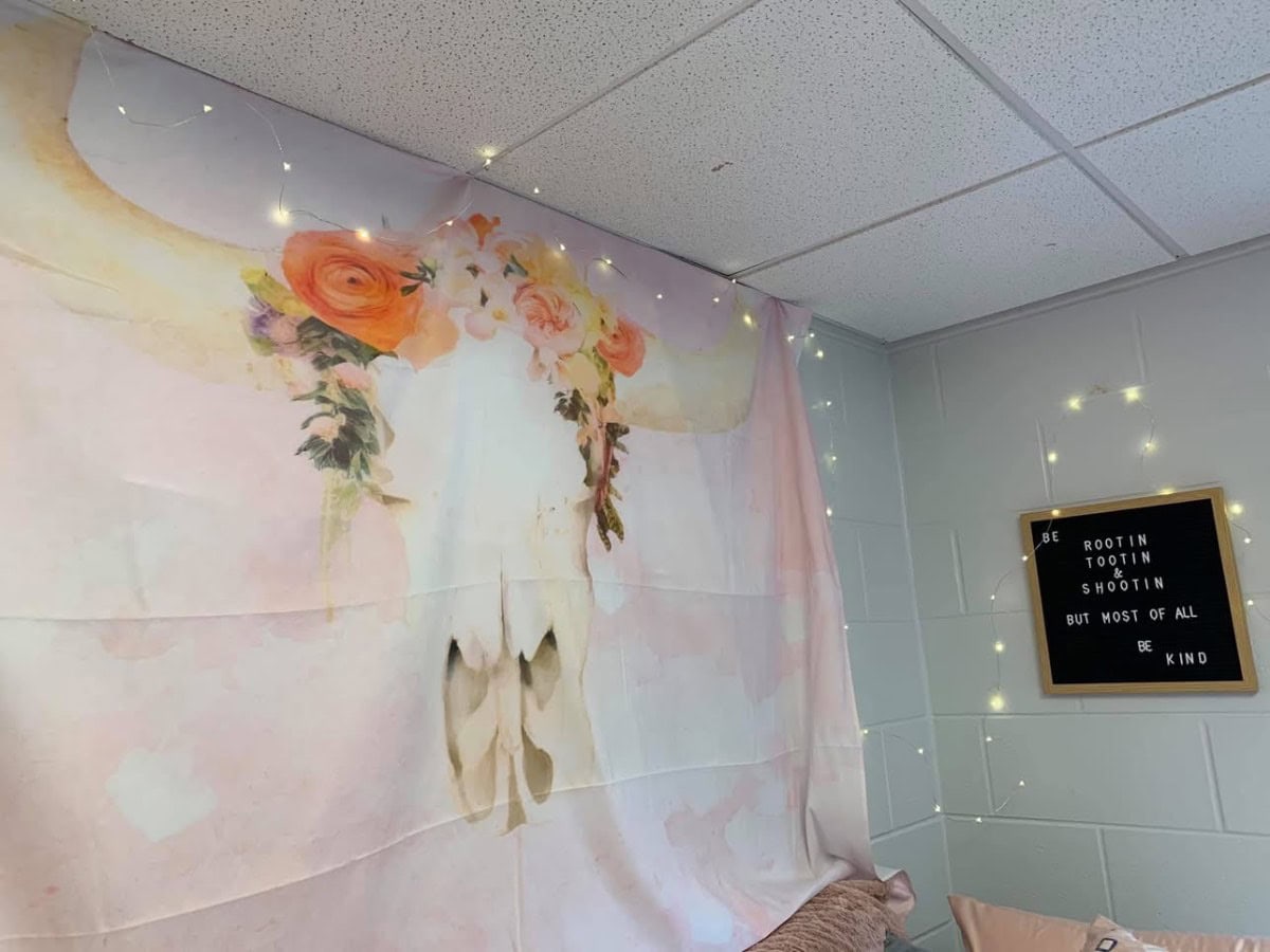 A wall tapestry featuring a cow skull adorned with flowers hangs in the corner. Nearby, a letter board displays, "Be rootin, tootin, shootin, but most of all, be kind," reminding one of a checklist for college freshmen. Fairy lights are draped around the area, creating a cozy ambiance.