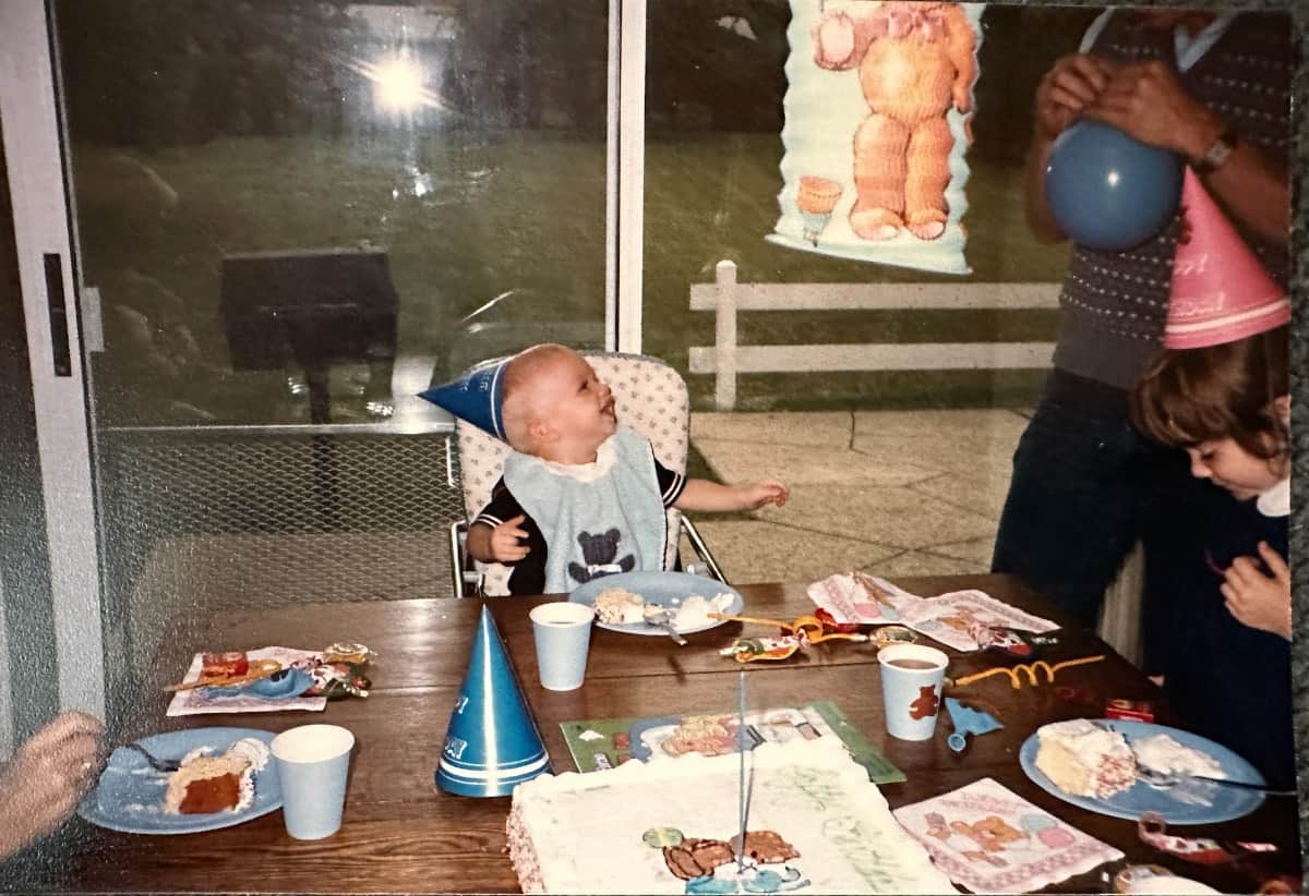 A baby wearing a blue party hat and bib sits in a high chair at a table with cake and party supplies, smiling up at an adult holding a balloon—a joyful scene sure to become a fond recollection. Another child leans on the table amid decorations and a window.