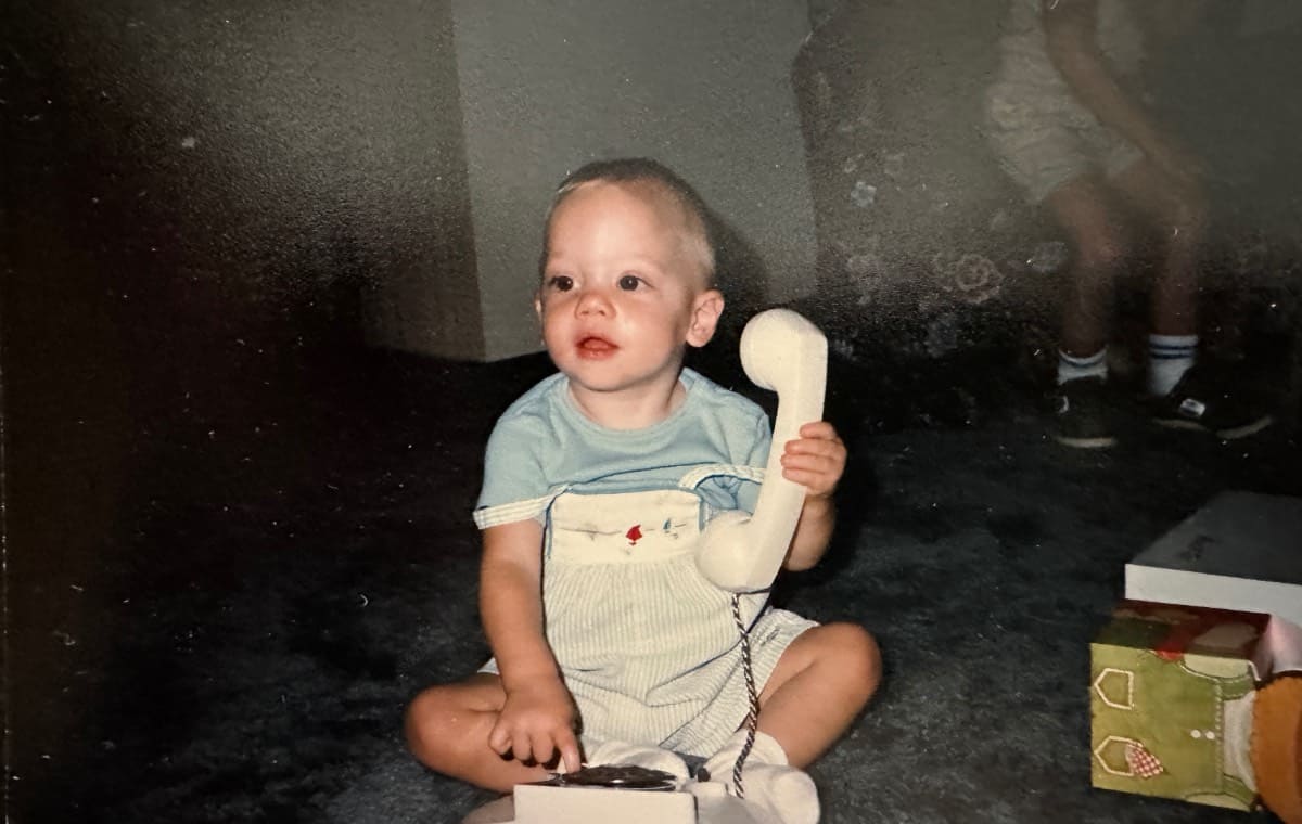 A toddler sits on a carpeted floor, holding a white telephone receiver and looking up in a moment of recollection. Dressed in light blue, the child is surrounded by presents, with another person's legs partly visible in the background.